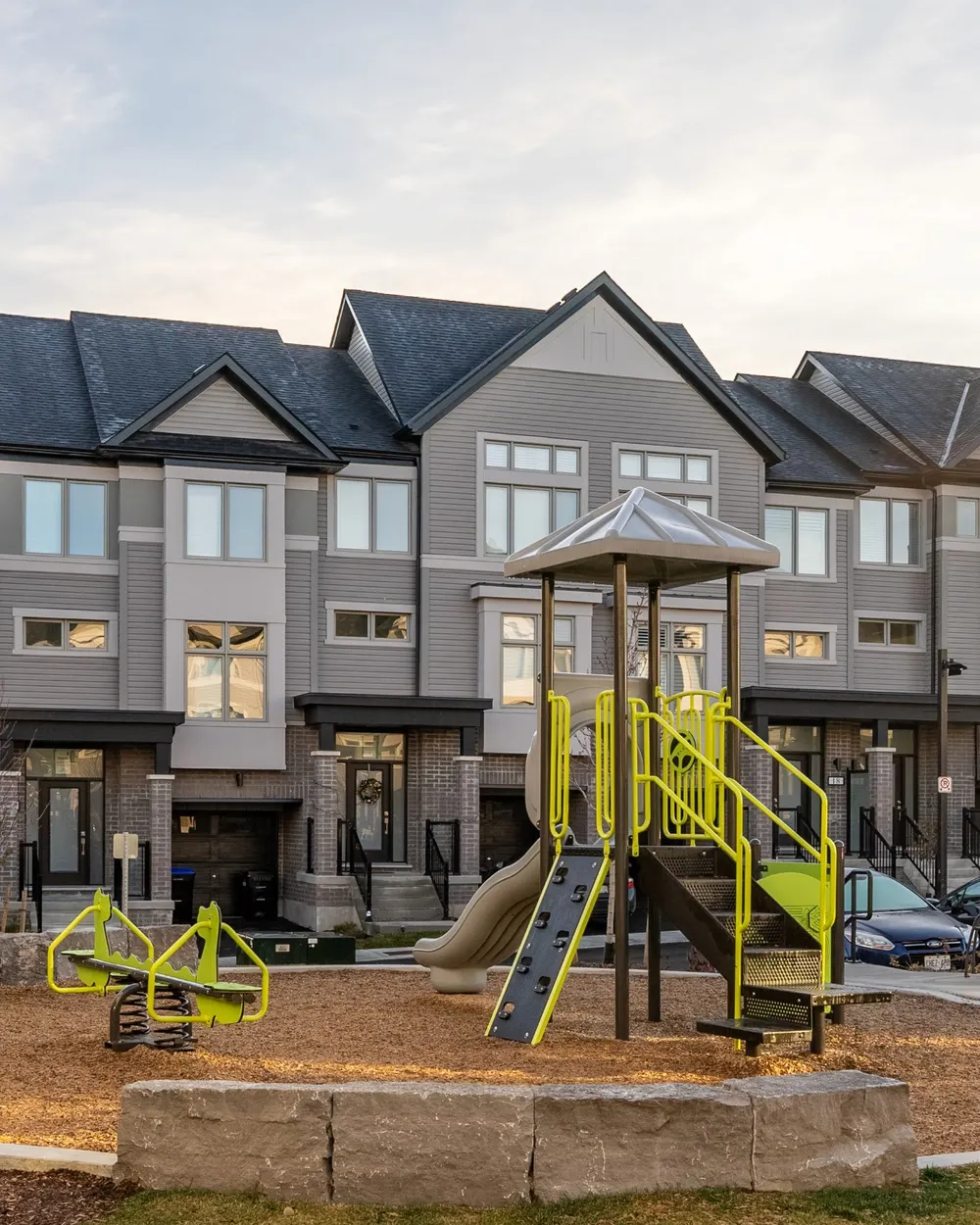 Three-storey back-to-back townhomes at The Townes at Deer Springs in Tottenham, Ontario, with grey and white siding, dark roofs, and a children's playground in the parkette in front